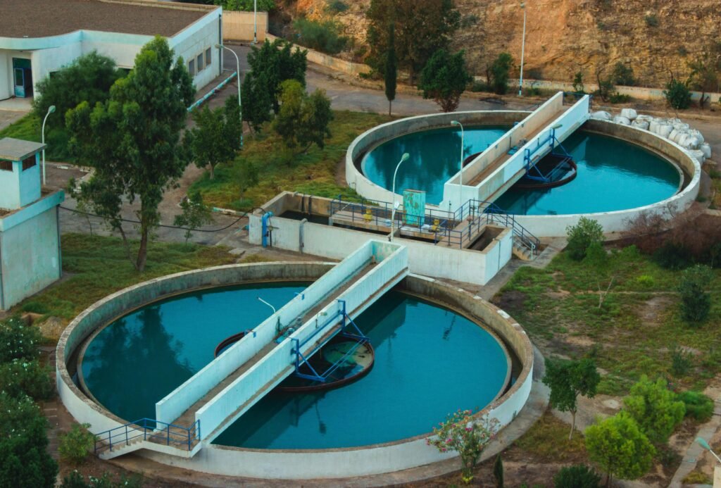 Aerial drone view of a modern sewage treatment plant featuring large circular settling ponds and interconnected basins amid green fields.