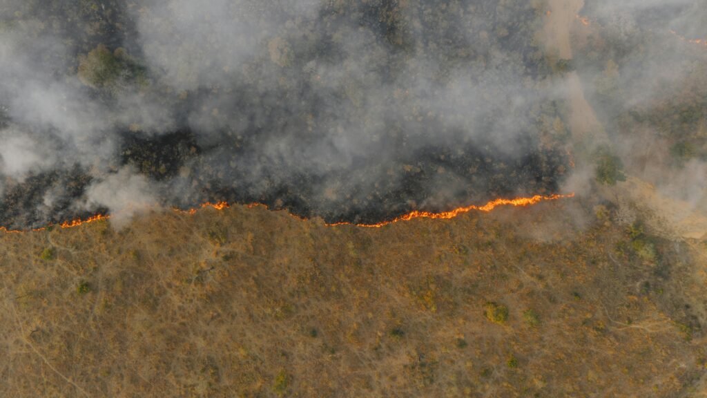 Aerial drone view captures a wildfire rapidly spreading flames across vast dry grassland under hazy skies, illustrating climate-driven blazes in the Southern Hemisphere.