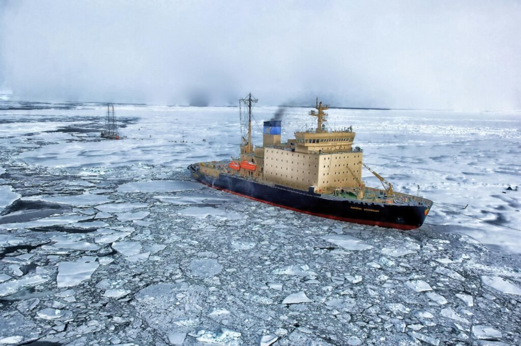 A dark gray ship sails through fragmented sea ice on calm Arctic waters under a cloudy sky.