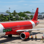 A modern red-and-white low-cost airline plane resembling AirAsia sits on a sunny airport tarmac while ground crew refuel it from a sustainable aviation fuel truck labeled 'SAF 100% Renewable'. Green solar panels cover nearby airport buildings amid lush greenery, with a digital dashboard on the plane showing 'CO2 Reduced by 80%' under a clear blue sky.